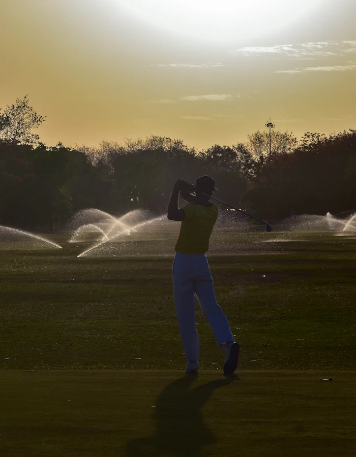 A golfer mid-swing as sprinklers run at golden hour